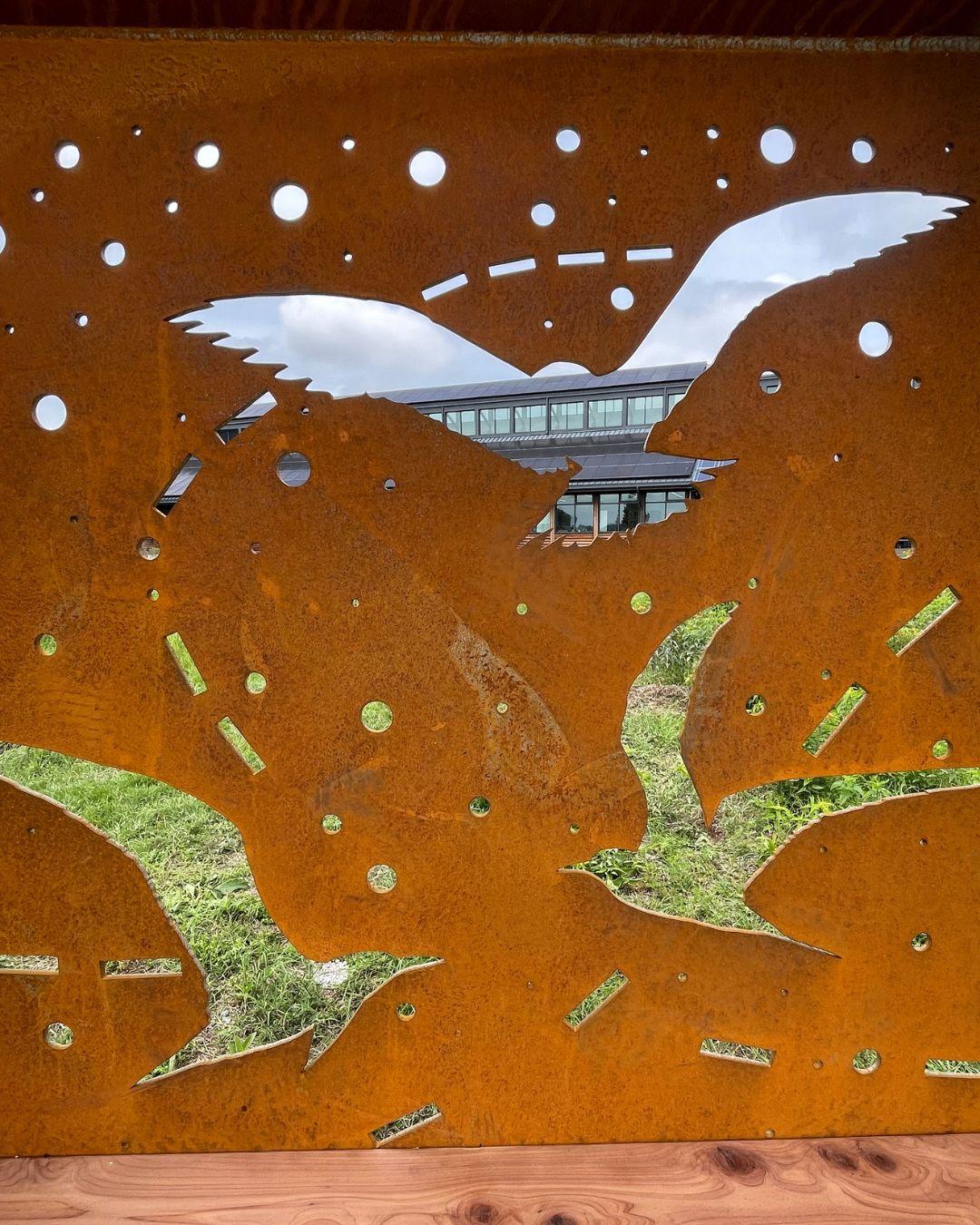 Photograph of a rusted metal screen with bird cutouts framing a building and garden.