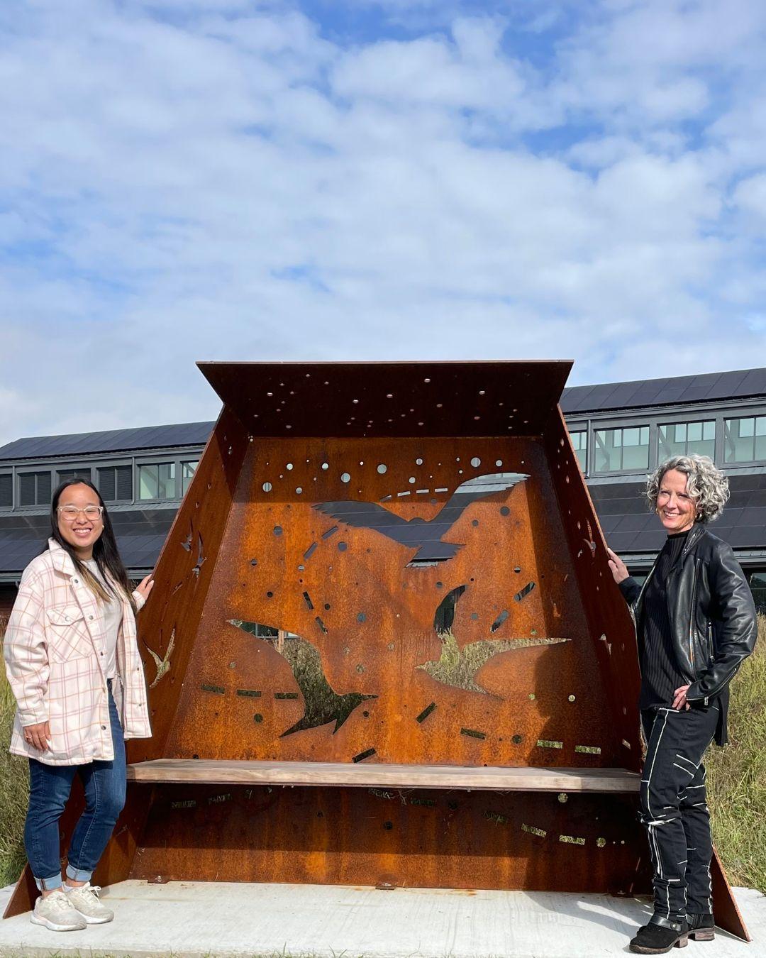 Katie Bui and Deirdre Murphy by a rusted metal sculpture with bird cutouts under a cloudy sky.