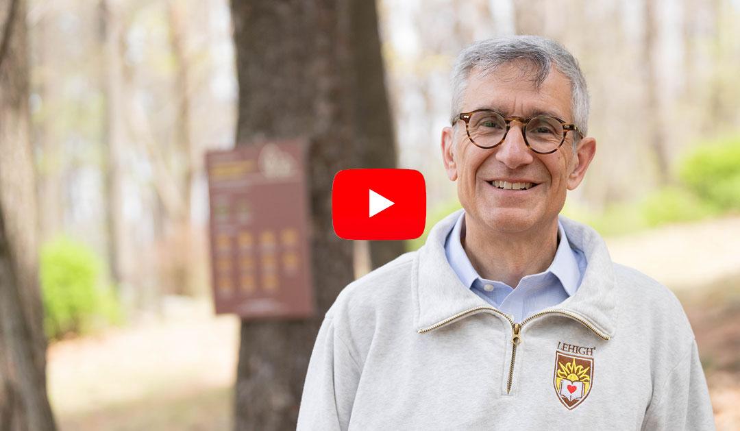 Photograph of a smiling man in a sweater outdoors with a play button overlay.