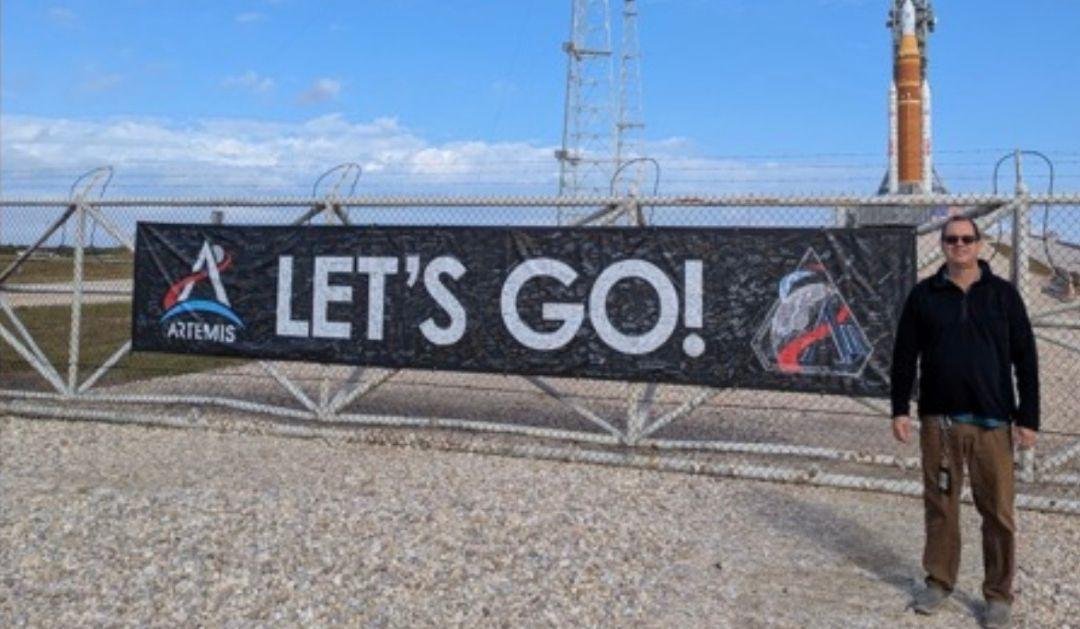 Photograph Mark Riddle by an Artemis "LET'S GO!" banner with a rocket in the background.