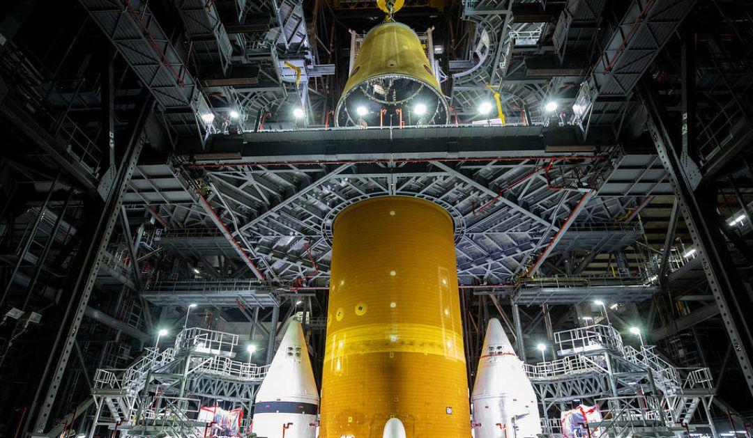 Photograph of a massive orange rocket core stage being assembled inside a hangar.