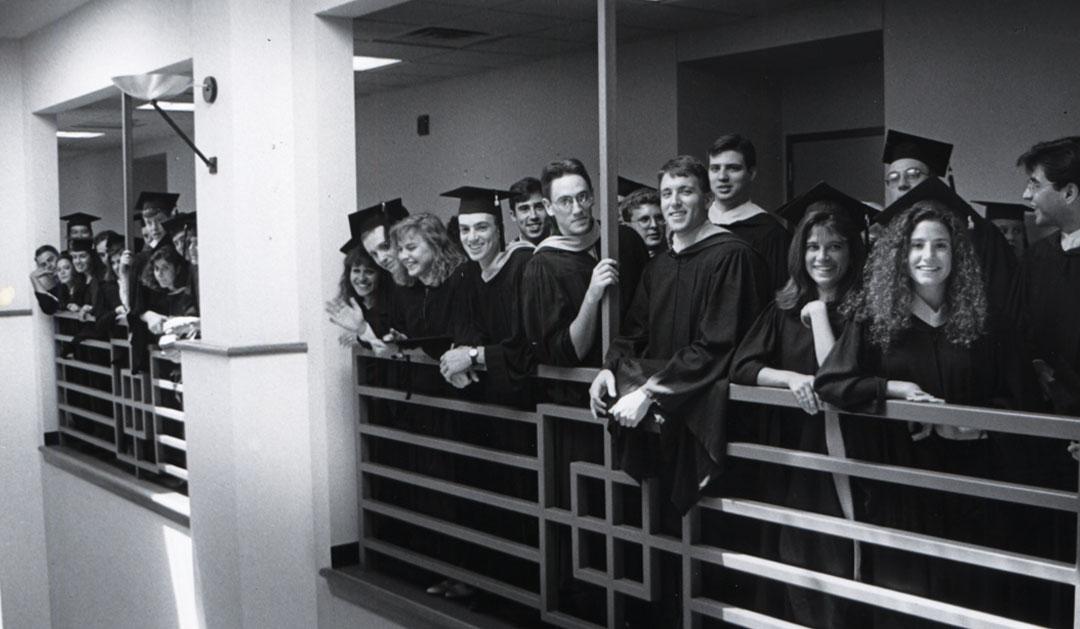 Black and white photograph of graduates in caps and gowns on an indoor balcony.
