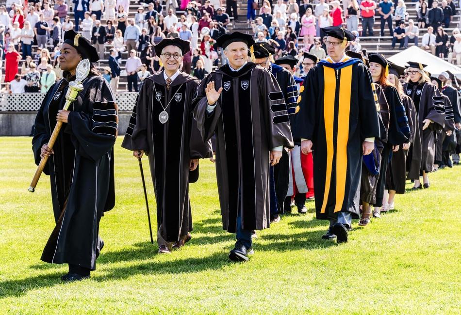 Photograph of university officials in academic regalia walking on a lawn at graduation.