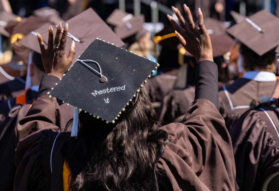 Photograph of a graduate in a brown cap and gown raising their hands at a graduation ceremony.