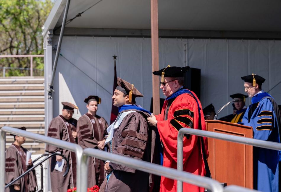 Photograph of a faculty member hooding a graduate in a brown gown on an outdoor stage.