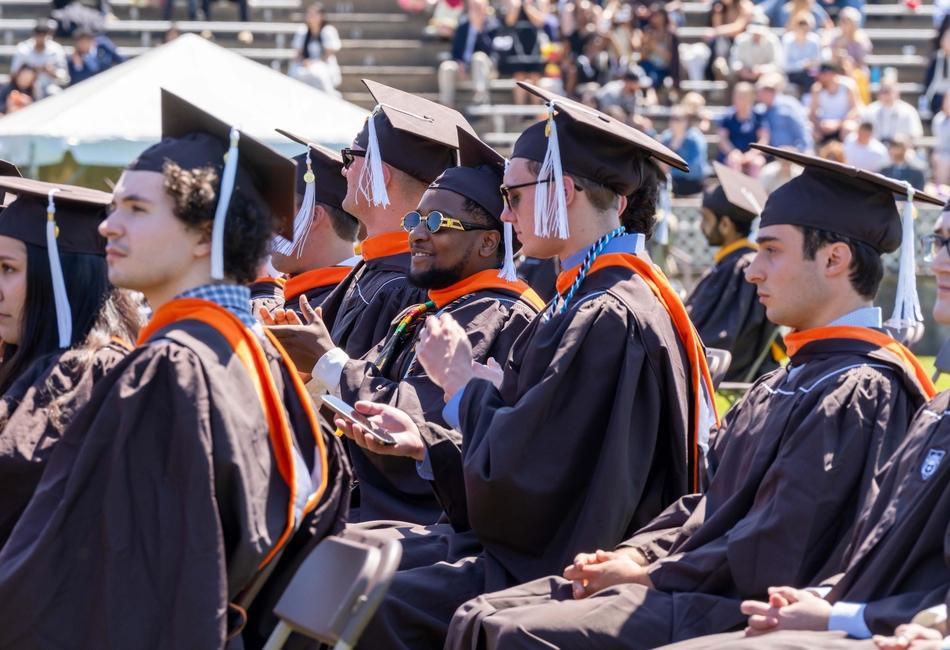 Photograph of graduates in brown caps and gowns seated at an outdoor graduation ceremony.