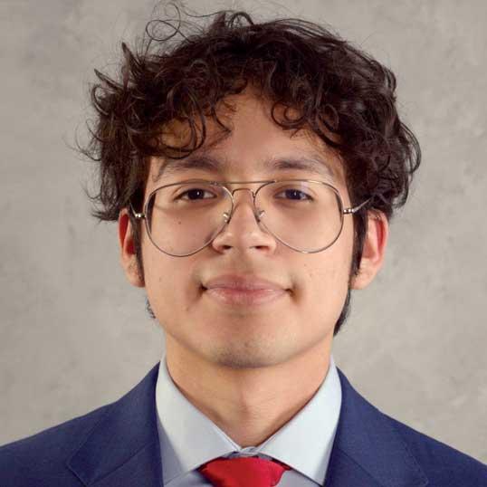 Headshot photograph of a man with curly hair and glasses wearing a blue suit and red tie.