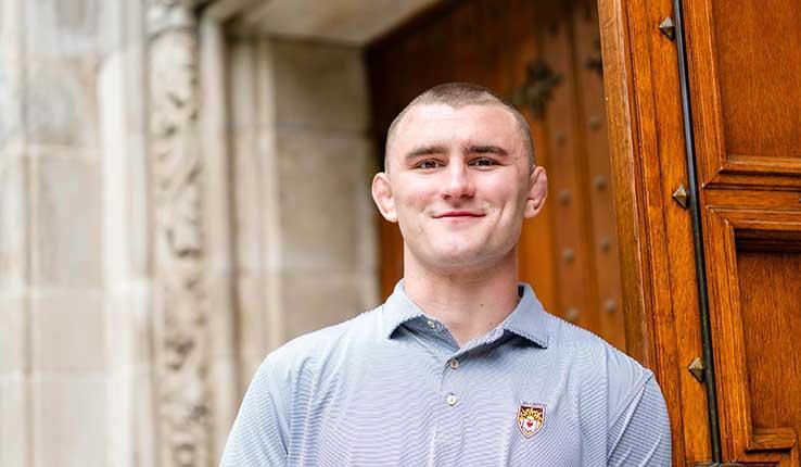 Photograph of smiling man in a light blue polo shirt in front of a stone building doorway.