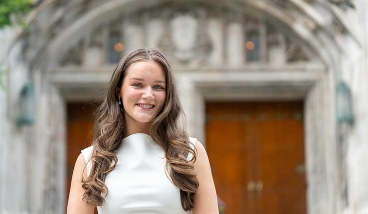 Photograph of a smiling woman in a white dress in front of an arched stone building.