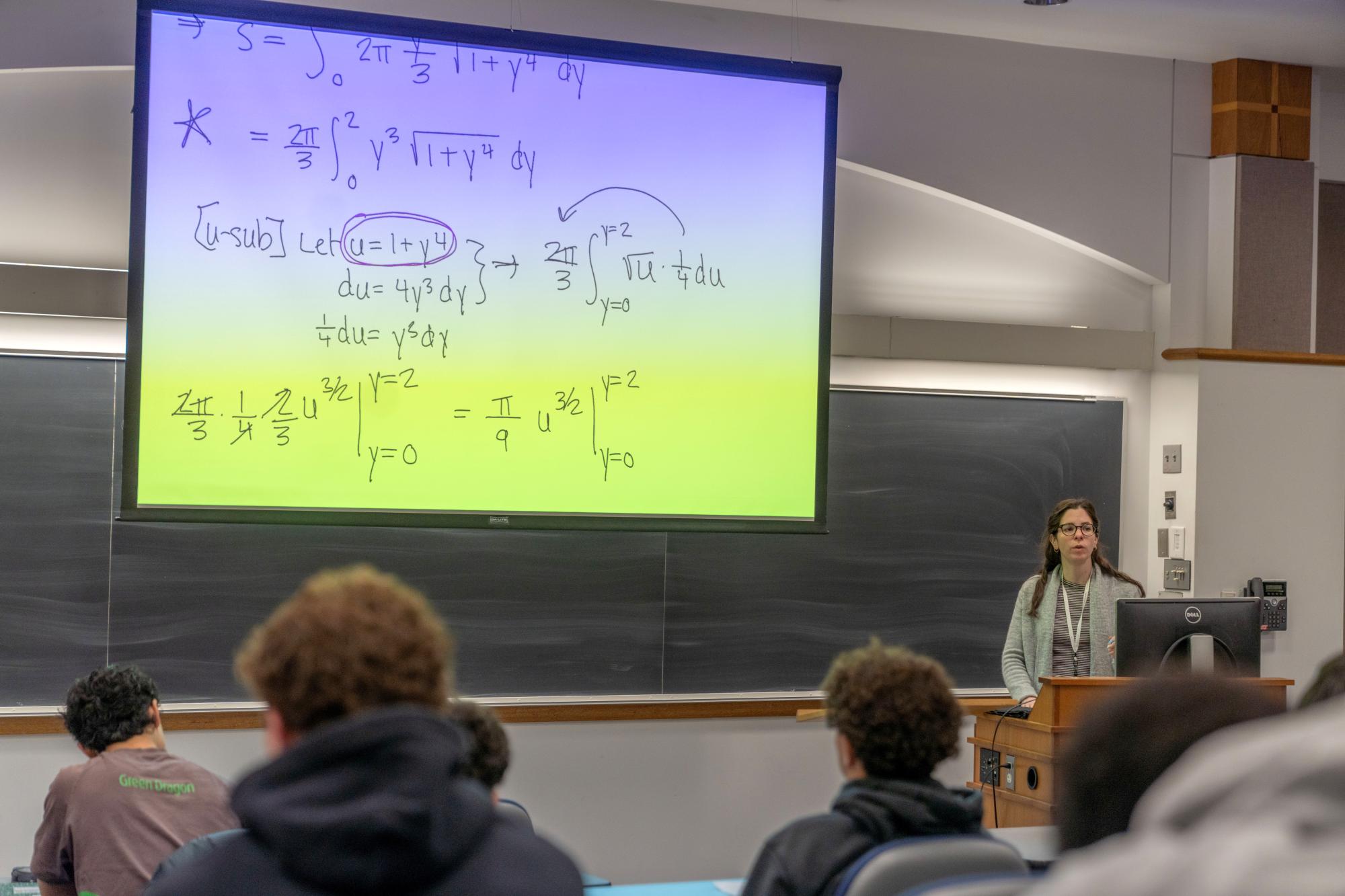 Photograph of a professor lecturing in a classroom with math equations on a large screen.