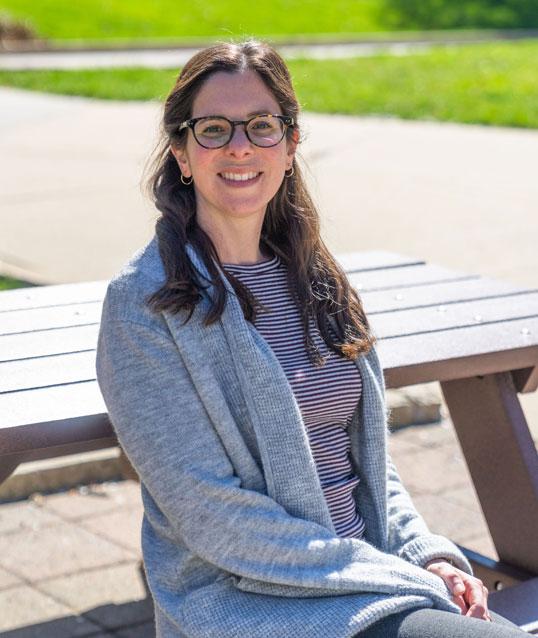 Photograph of a smiling woman sitting at an outdoor picnic table.