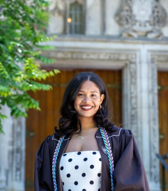 Photograph of a smiling graduate in regalia in front of a stone university building.