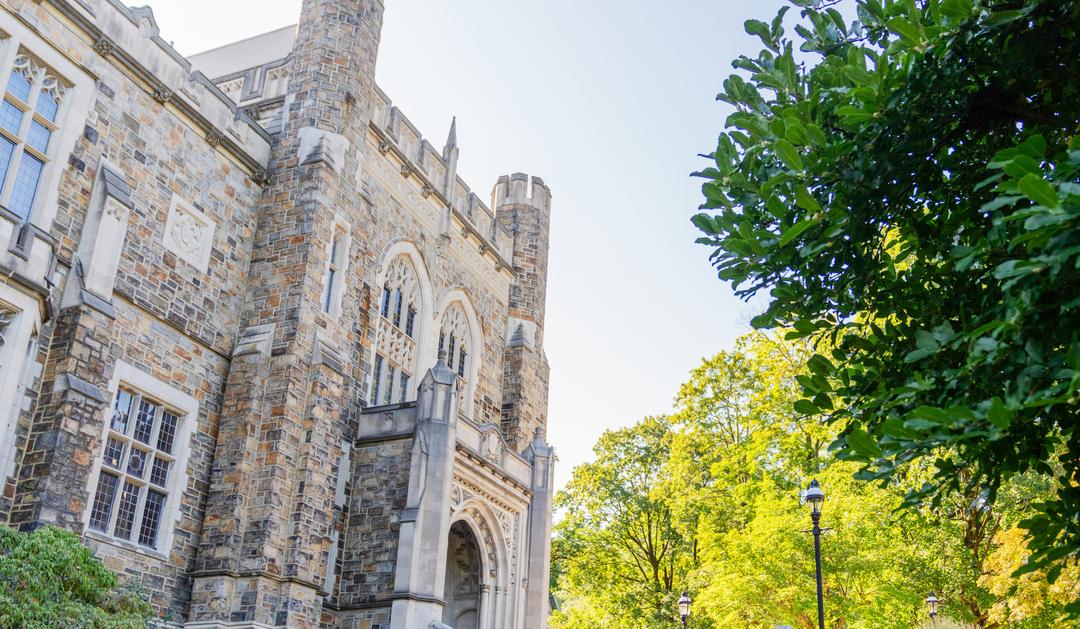 Photograph of a collegiate gothic stone building framed by lush green trees.