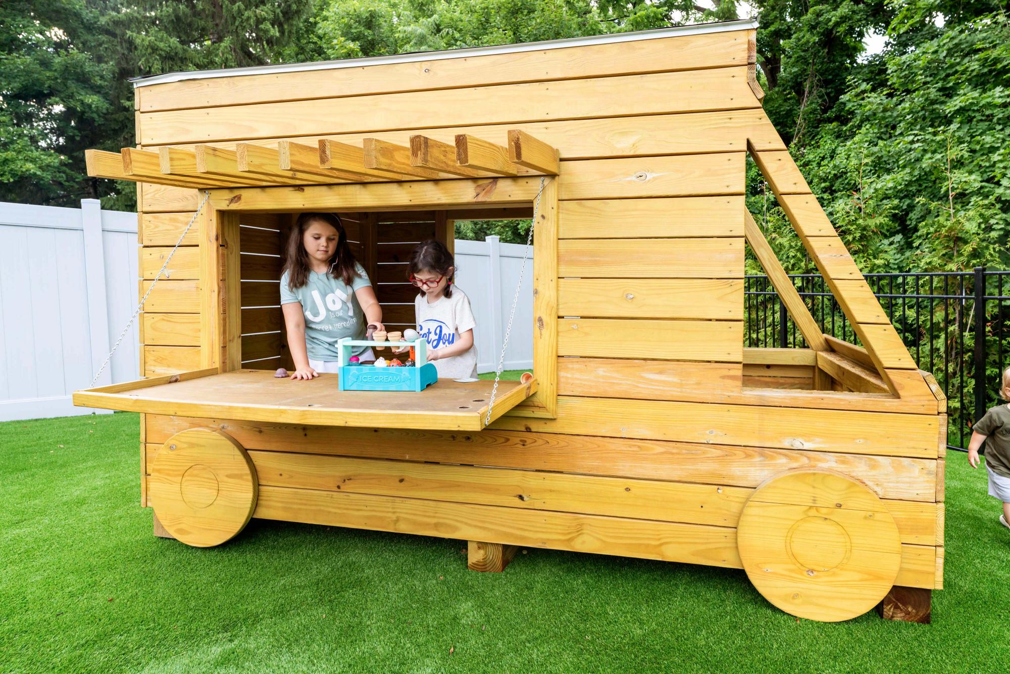 Two children play in a wooden truck-shaped playhouse in a backyard.