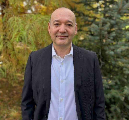 Smiling man in a dark blazer and white shirt with autumn foliage in the background.