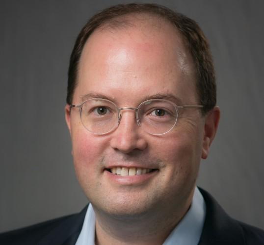 Professional headshot photograph of a smiling man wearing glasses.