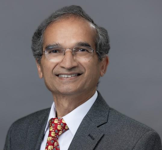 Photograph of a smiling man wearing glasses, a grey suit, and a red elephant-patterned tie.