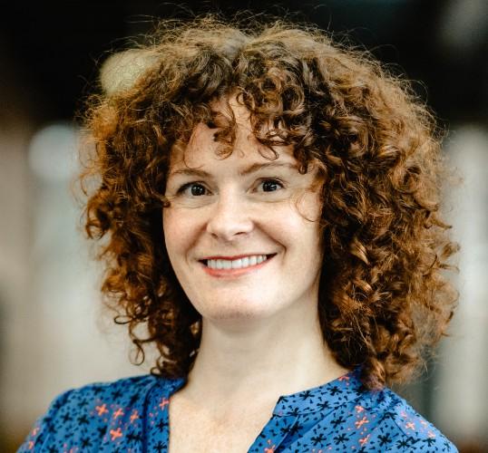 Photograph of a smiling woman with curly auburn hair wearing a blue patterned shirt.