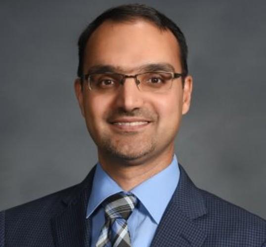 Headshot of a smiling man with glasses, wearing a blue shirt and patterned tie.