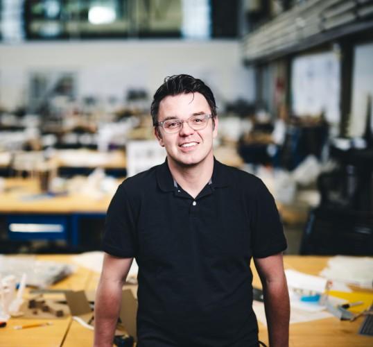 Photograph of a smiling man in a black polo shirt and glasses in a design workshop.