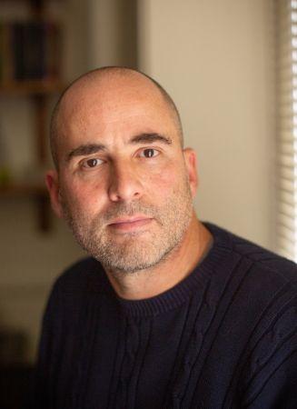 Portrait photograph of a man with a shaved head and stubble wearing a navy sweater.