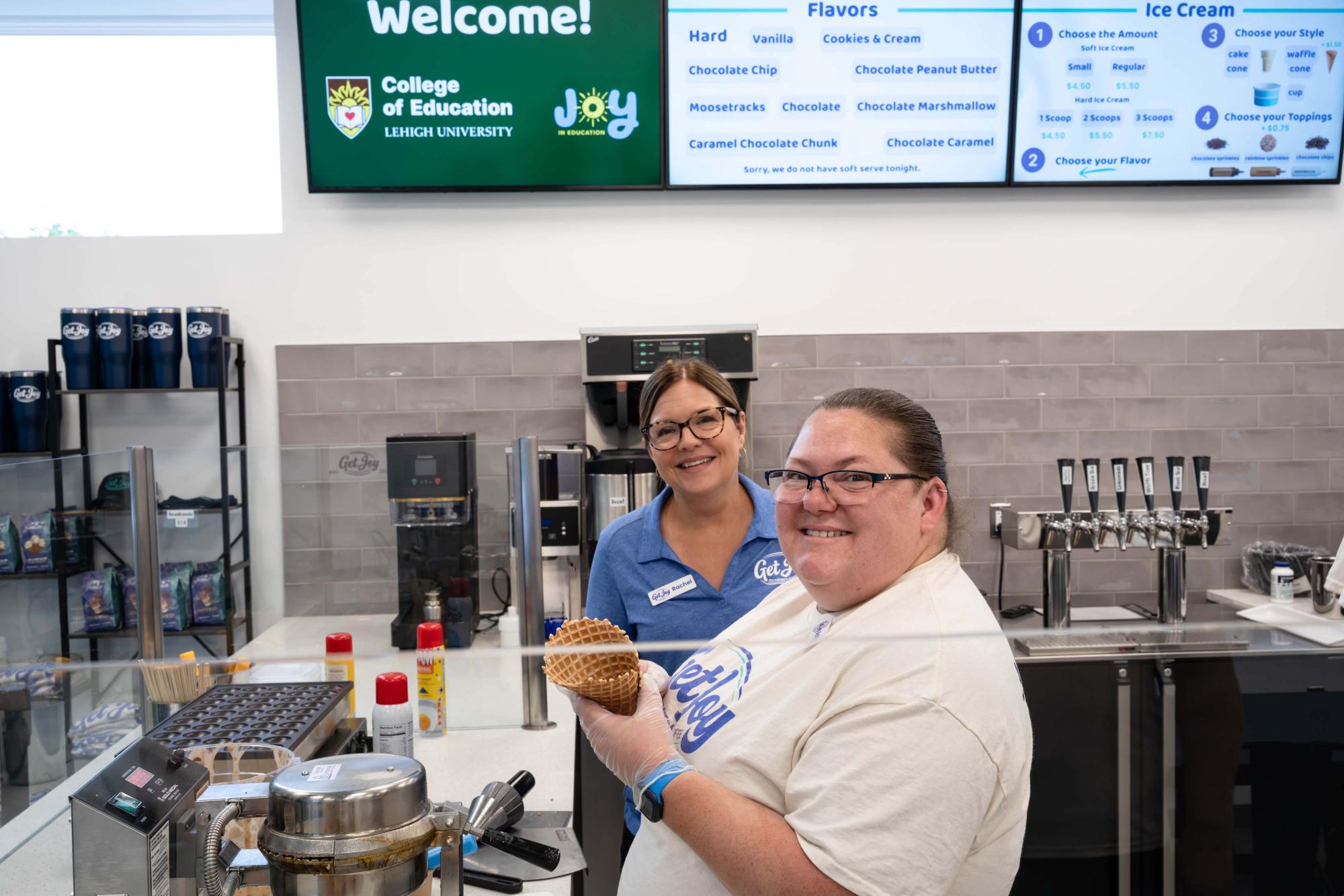 Photograph of two women smiling behind an ice cream counter, one holding a cone.