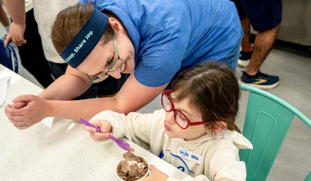 Photograph of a woman watching a young girl in red glasses eat chocolate ice cream.