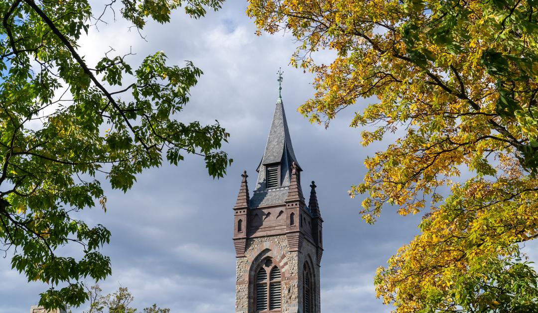 Photograph of a Gothic stone spire framed by colorful leaves and a cloudy sky.