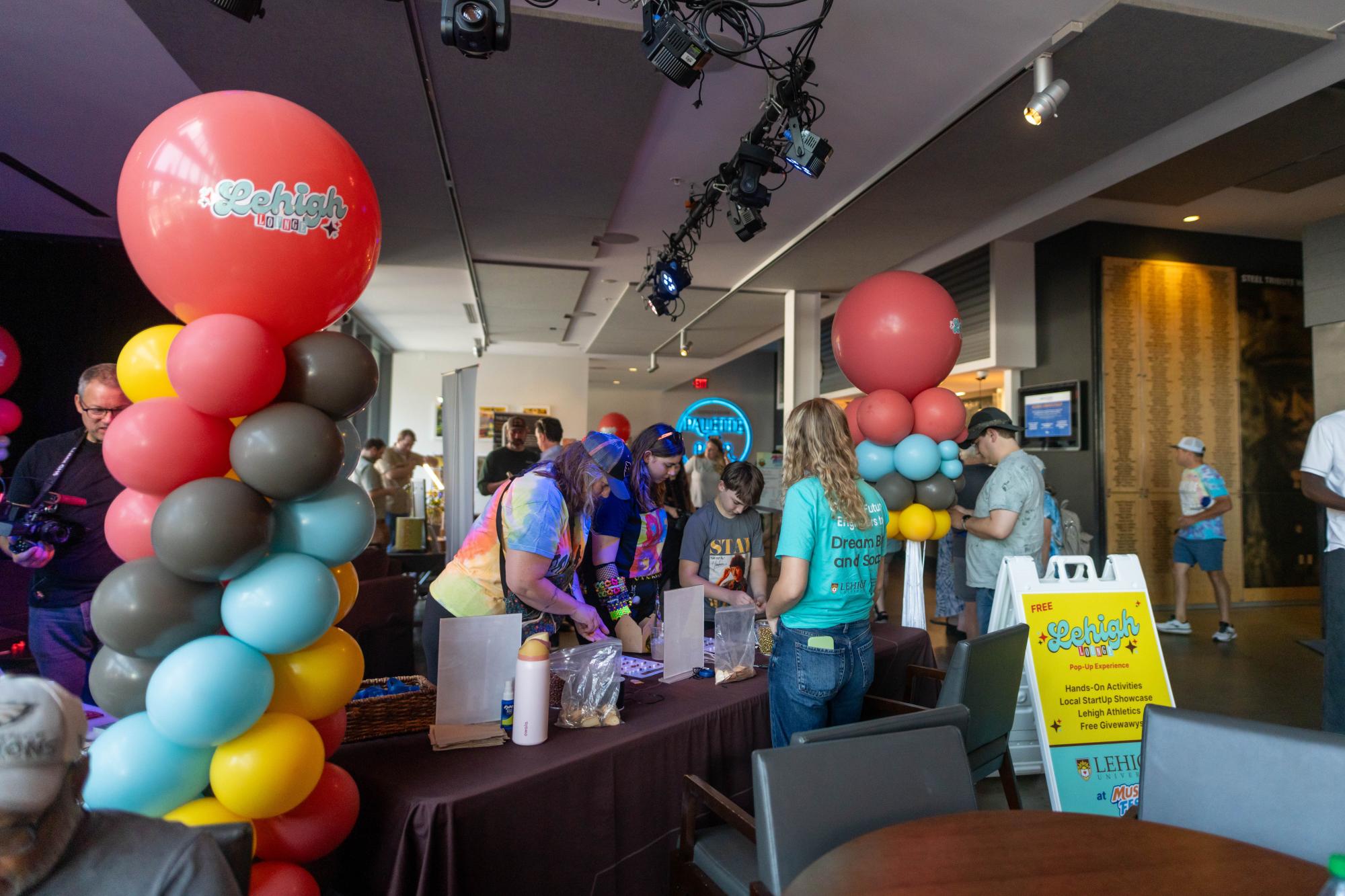 Photograph of a festive event with colorful balloon columns and people at activity tables.