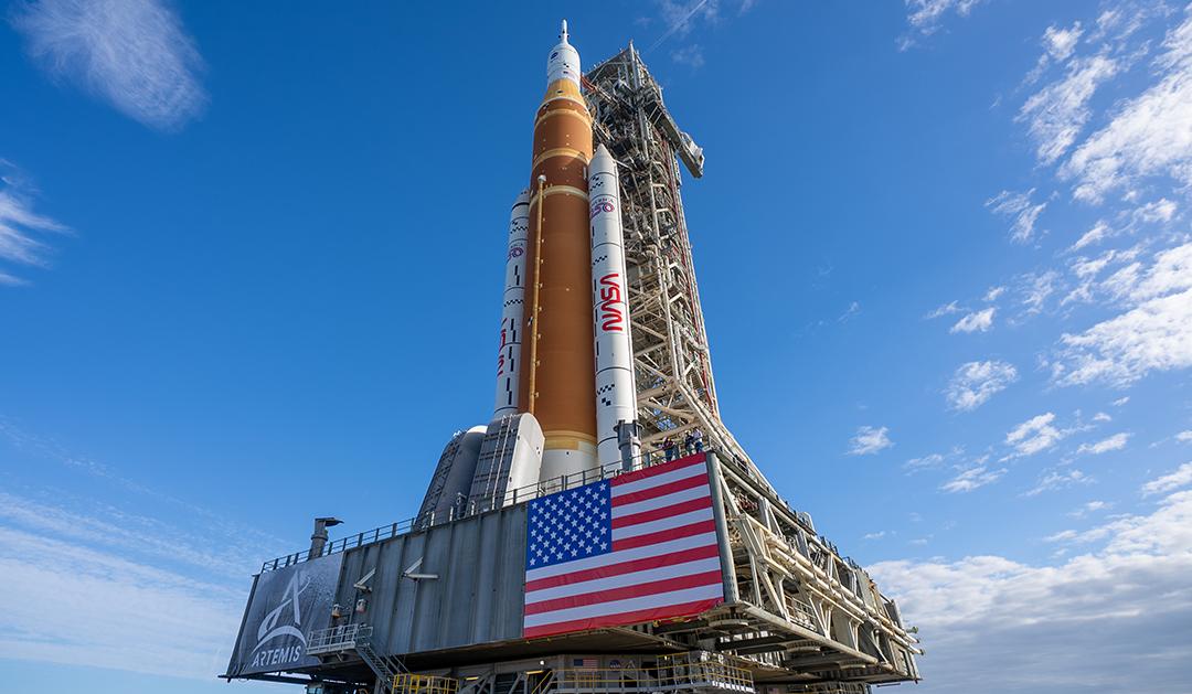 Photograph of NASA’s Artemis rocket on its launch pad with a large American flag.