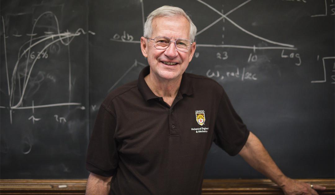 Photograph of a smiling male professor in front of a blackboard with mathematical diagrams.