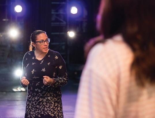 Woman in glasses gesturing with clenched fists on a stage with bright spotlights.
