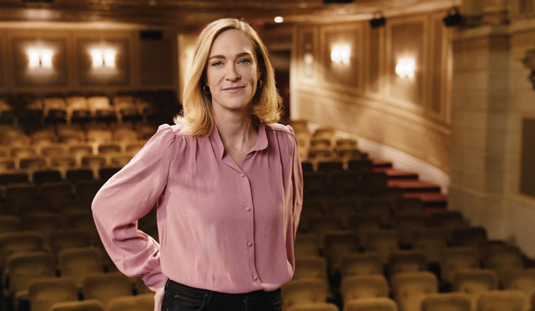 Photograph of a smiling woman in a pink blouse in an empty theater auditorium.