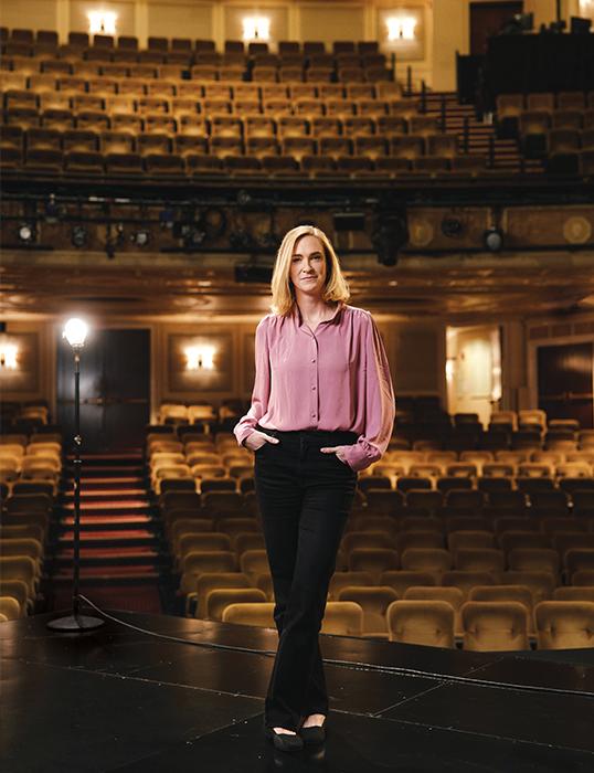 Photograph of a woman in a pink shirt standing on an empty theater stage.