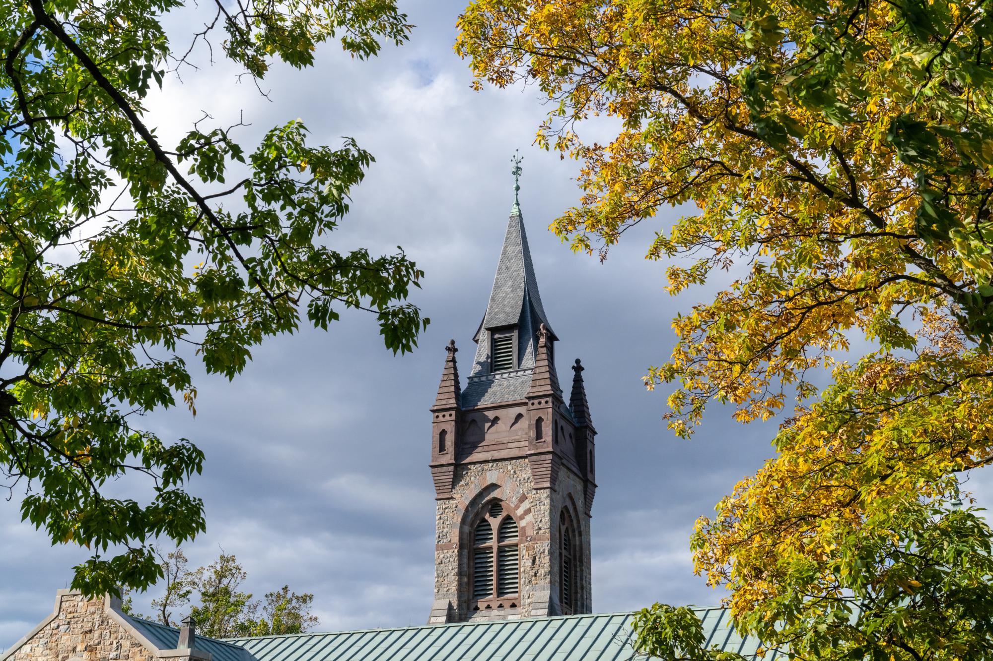 Photograph of a stone Gothic tower framed by yellow autumn leaves against a cloudy sky.
