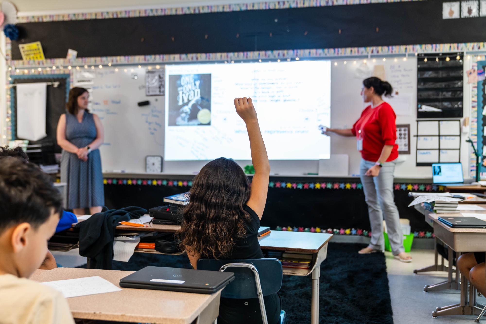 A teacher points to a projector screen as a student raises her hand in a classroom.