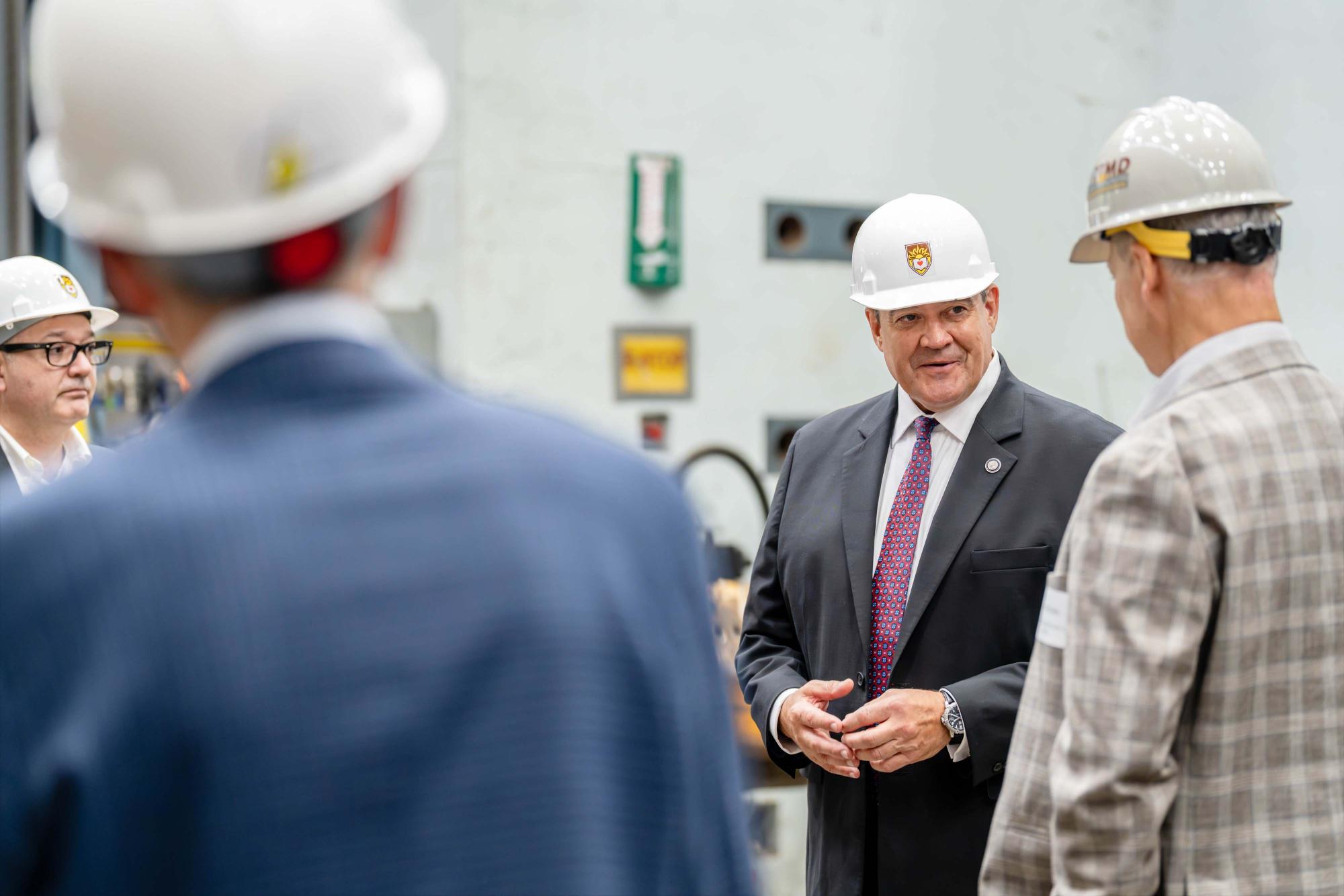 Photograph of a man in a suit and hard hat speaking to others in an industrial setting.