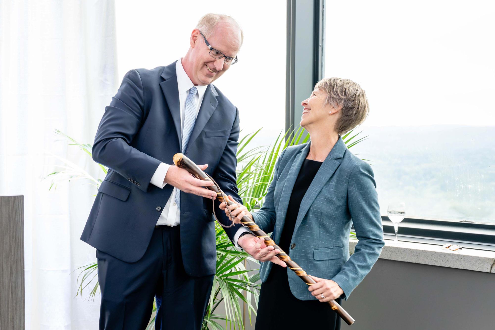 Photograph of a man in a suit presenting a wooden flute to a smiling colleague.