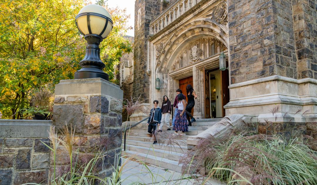 Students walking down the steps of a stone collegiate building in autumn.