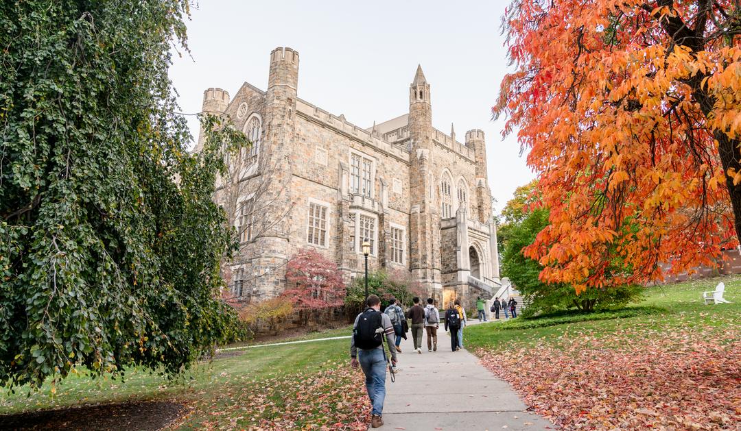 Photograph of students walking toward a stone collegiate building among autumn foliage.