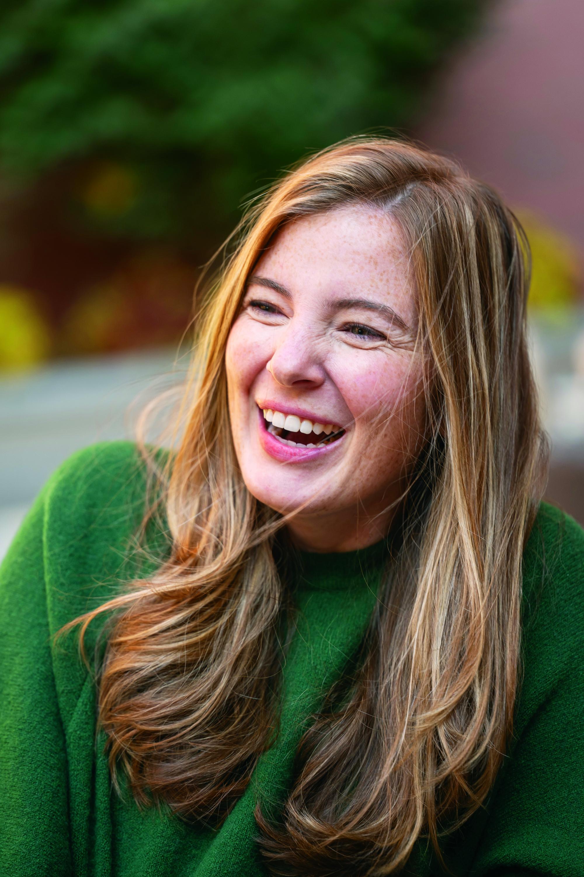 Photograph of a woman with long brown hair laughing, wearing a green sweater.