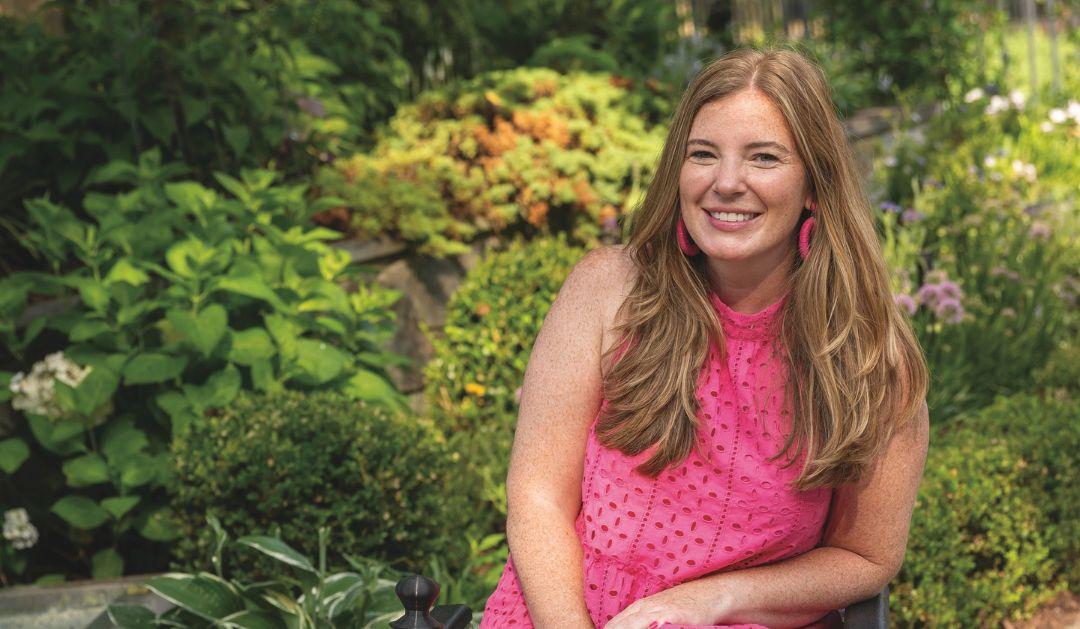 Photograph of a smiling woman in a bright pink top sitting in a lush garden.