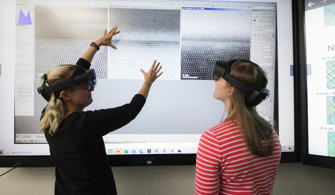 Two women wear VR headsets in front of a large touchscreen