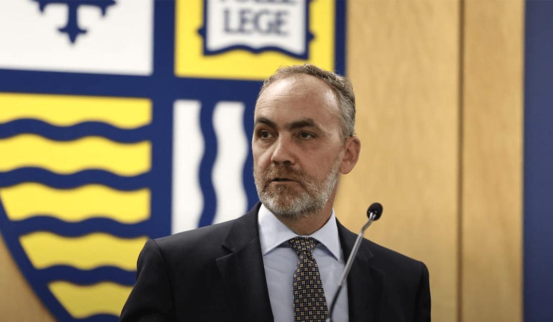 Jeremy Gibson in a suit and tie speaking at a podium, with a microphone in front of him.