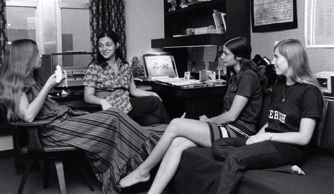 Four students chat at a desk in a dorm room