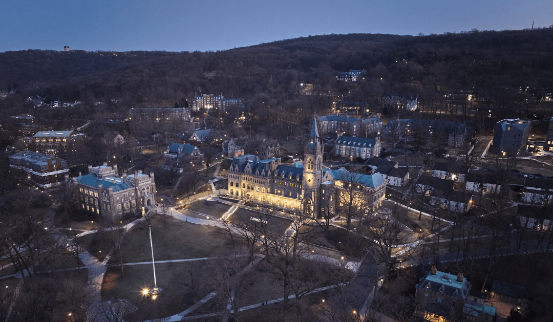 Aerial view of a campus at twilight, buildings illuminated, surrounded by trees and hills.