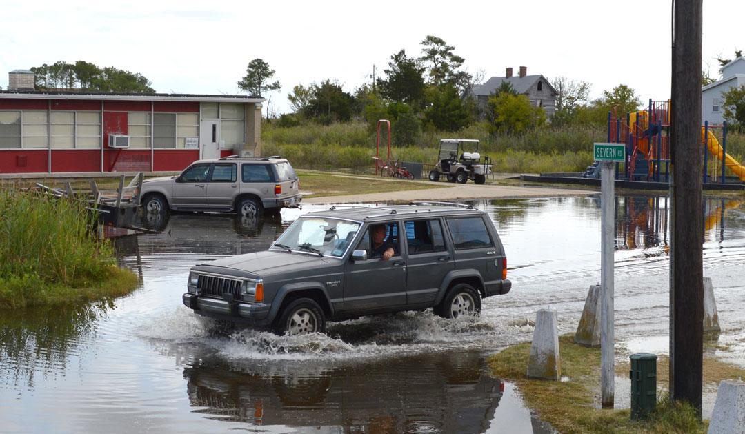 A gray SUV drives through a flooded road near a playground and buildings.