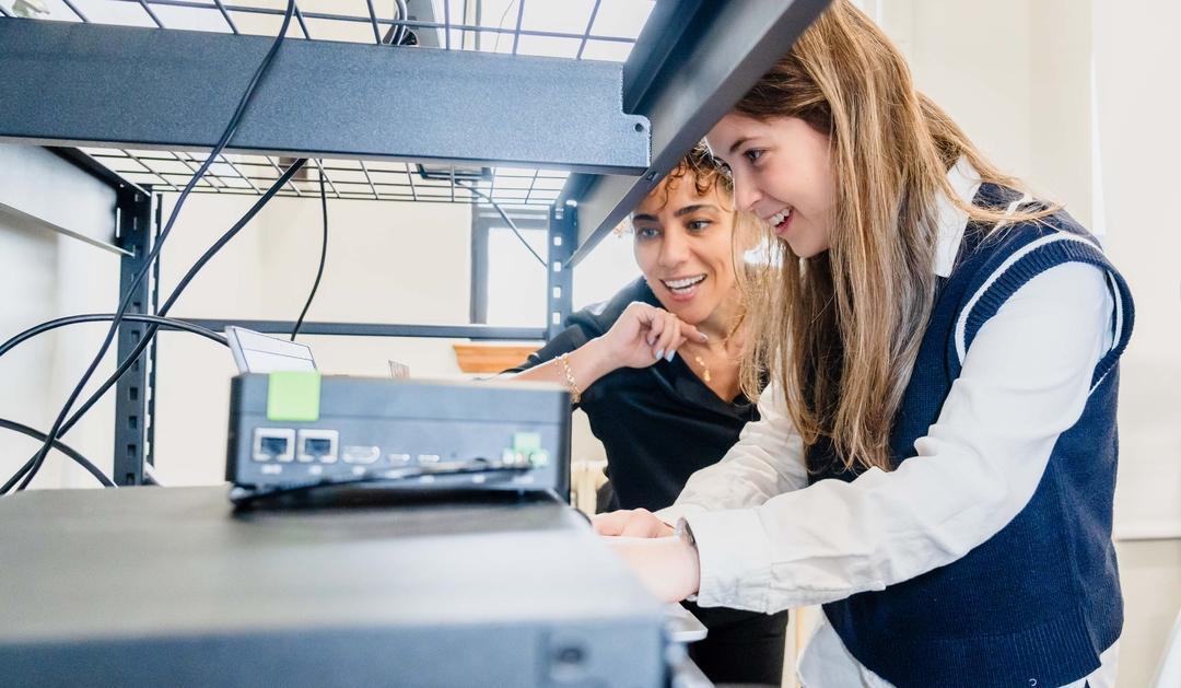 Two smiling women inspecting network equipment on a shelf. 