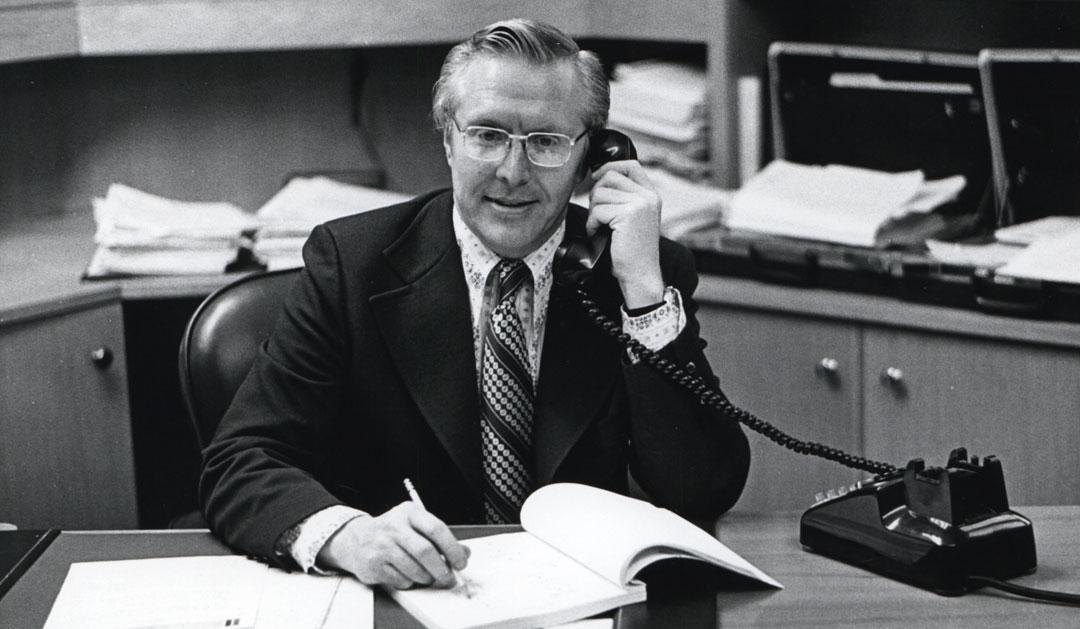 Black and white photograph: Man in suit and glasses talks on phone, writes at office desk.