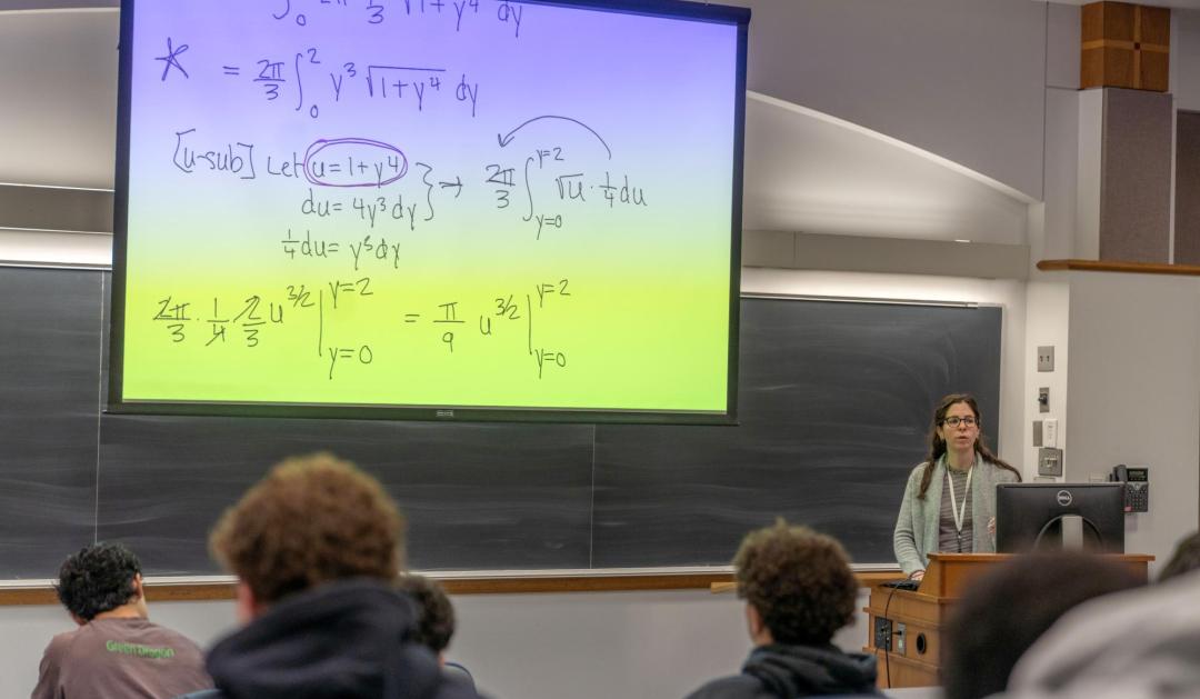 Photograph of a professor lecturing in a classroom with math equations on a large screen.